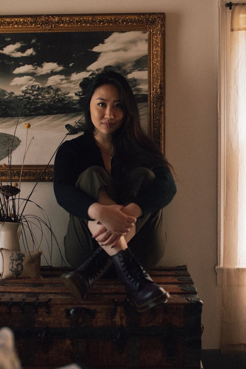 Michelle Saya seated on an old travelling trunk, a gilt-framed painting behind her